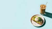 Avocado toast with poached egg and microgreens on plate, iced coffee beverage, minimalist flat lay on light blue background.