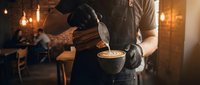 Professional barista in black gloves pouring steamed milk creating latte art in ceramic cup at cozy coffee shop with warm ambient lighting and rustic interior.
