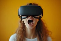 Young woman with curly hair wearing virtual reality headset expressing excitement with open mouth against vibrant yellow background.