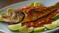 Grilled whole fish with spicy tomato sauce and lime wedges on white plate. Fresh seafood dish with herbs and citrus garnish for restaurant menu design.