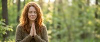 Peaceful redhead woman meditating with prayer hands in serene forest setting during golden hour light for wellness and mindfulness practices.
