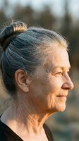 Senior woman with gray hair in top knot, smiling in profile outdoors during golden hour. Mature female with lined face and natural aging features in natural light.