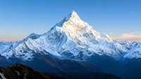 Majestic snow-covered mountain peak rising above mountain range at sunrise, dramatic alpine landscape with sunlit summit against clear blue sky and misty valleys.