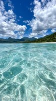 Crystal clear turquoise water with rippling patterns under dramatic cloudy sky, tropical mountains and lush green coastline creating paradise seascape for travel content.
