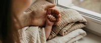 Adult hand gently holding tiny newborn fingers on soft linen blankets near a window, capturing tender family bond and new life.