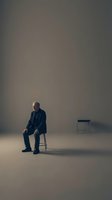 Middle-aged man in dark business suit sitting on chair in minimalist studio setting with empty space, conveying solitude, waiting, or contemplation for corporate concepts.