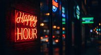 Neon Happy Hour sign glowing red in urban nightlife district with colorful bokeh lights and blurred city background creating atmospheric evening scene.