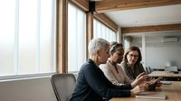 Mature professionals reviewing digital tablet together in bright office space with wooden beams and large windows, suitable for business, teamwork, and corporate training materials.
