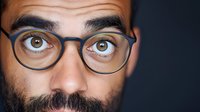 Close up portrait of young Middle Eastern man with glasses and beard looking directly at camera with intense brown eyes against dark background.