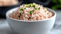 Steamed pink rice garnished with fresh chopped chives in white ceramic bowl on light background, close up view of healthy Asian cuisine side dish.