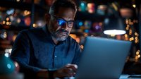 Middle-aged bearded man in glasses working late on laptop with blue light reflection, dark moody atmosphere with bokeh lights in background.
