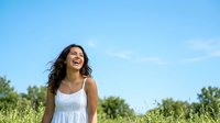 Young woman with long dark wavy hair and South Asian ethnicity wearing white sleeveless dress, smiling joyfully in sunny green field under blue sky.