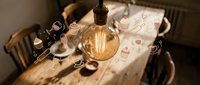 Vintage Edison bulb illuminates cozy workspace with retro camera, coffee cups and scattered papers on wooden table creating warm atmospheric lighting.