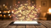 Digital tablet displaying glowing email icons with sparkles and light effects on wooden table in cozy home interior for communication technology concepts.