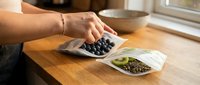 Person with medium skin tone reaching for fresh blueberries on wooden table during meal preparation, with healthy food ingredients and bowl nearby.