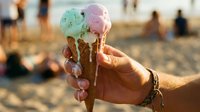Colorful ice cream cones held in hand at beach during golden hour sunset, perfect for summer food and lifestyle marketing campaigns.