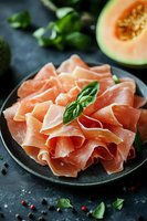 Sliced prosciutto ham with fresh basil and cantaloupe melon on dark plate for gourmet appetizer and charcuterie board presentation.