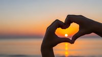 Hands forming heart shape silhouette against golden sunset over calm ocean water creating romantic peaceful moment for love wellness concepts.