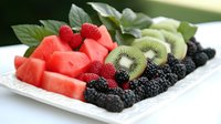 Fresh summer fruit platter with watermelon chunks, ripe blackberries, juicy raspberries and sliced kiwi arranged on white plate with green leaves, ready for healthy snack.