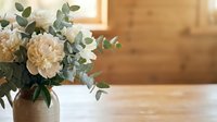 Ceramic vase with white peonies and eucalyptus on a wooden table in a warm rustic interior with soft natural light.