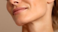 Close-up of woman's lips and jawline against neutral beige background, showcasing natural beauty and facial features for skincare or cosmetic advertising.