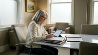 Mature woman with gray hair and glasses, wearing a beige cardigan, working at a desk with documents and laptop in a bright office setting.