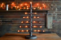 Modern industrial pipe lamp with orange glowing bulbs arranged in minimalist tree shape on wooden table against brick wall with festive holiday lighting.