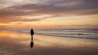 Silhouetted figure standing alone on sandy beach at golden hour sunset with ocean waves and dramatic sky, suitable for travel and lifestyle content.