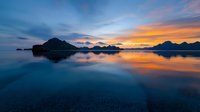 Dramatic sunset over tranquil fjord waters with silhouetted mountain peaks and vibrant orange sky reflecting on calm surface, creating mirror-like symmetry in nature.
