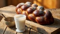 Fresh braided challah bread with sesame seeds on wooden cutting board alongside glass of milk, traditional Jewish bakery product for Shabbat celebration.
