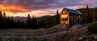 Wooden cabin glowing warmly at sunset in remote mountain wilderness surrounded by evergreen forest and rocky terrain during golden hour.