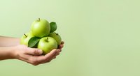 Hand holding three fresh green apples with leaves against pale yellow-green background, representing healthy eating, nutrition, organic produce, and wellness for food and lifestyle marketing.
