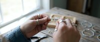 Skilled artisan hands crafting delicate wire jewelry with precision tools on wooden workspace near bright window light for handmade accessories creation.