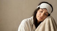 Young Asian woman wearing white sleep mask resting peacefully with soft blanket in cozy bedroom setting for relaxation and wellness content.
