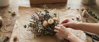 Florist arranging dried flowers and baby's breath into a rustic bouquet on kraft paper workspace with twine and greenery for wedding or event decoration.