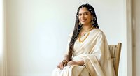 Young woman with long braided hair and South Asian ethnicity wearing cream-colored traditional garment, seated on wooden chair against neutral background. Suitable for cultural fashion, lifestyle, and portrait projects.
