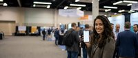 Young Hispanic businesswoman holding smartphone at corporate conference center with blurred crowd of professionals in background during networking event.