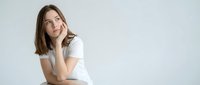 Young Caucasian woman in white t-shirt sitting with hand on chin in thoughtful pose against neutral background for lifestyle and wellness concepts.