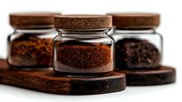 Ground coffee in glass jars with cork lids on wooden coasters against white background, selective focus on center jar showing fresh coffee granules texture.