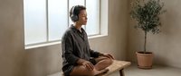 Young Asian woman meditating peacefully in minimalist room with natural light, sitting cross-legged on wooden bench wearing casual clothing for wellness lifestyle.