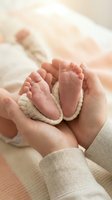 Adult hands cradling newborn infant feet wrapped in cream knit booties, soft focus background with sleeping baby, maternity and parenting concept.