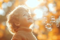Young caucasian child with curly hair in turtleneck sweater looking up at soap bubbles during golden sunset, backlit with bokeh autumn background and magical atmosphere.
