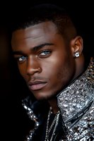 Young African American male model with striking blue eyes wearing silver metallic jacket and diamond earring against dark background, dramatic studio lighting.