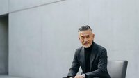 Mature male professional with gray hair and beard wearing dark blazer and glasses, seated in modern office environment with white walls, suitable for corporate, business, and leadership concepts.
