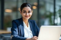 Professional South Asian businesswoman smiling at laptop in modern office environment for corporate presentations and marketing materials.