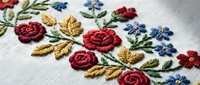 Delicate hand embroidered floral pattern with red roses, blue flowers and golden leaves on white fabric. Traditional needlework craft for textile design.