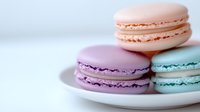 Pastel colored French macarons in pink, purple and turquoise arranged on white plate against light background, soft focus food photography for bakery marketing.