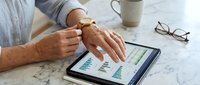 Senior businessman analyzing financial data charts and graphs on digital tablet at marble desk with coffee and glasses nearby.