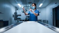 Female surgeon in blue scrubs and mask preparing medical syringe in modern operating room. Healthcare professional administering injection in sterile hospital environment.