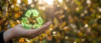 Caucasian hand holding glowing green recycling symbol against blurred autumn foliage background, representing environmental sustainability and eco-friendly practices for conservation campaigns.